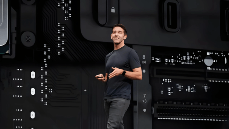 Un hombre sonriente de pie en un escenario con un fondo tecnológico, sosteniendo un dispositivo en su mano.