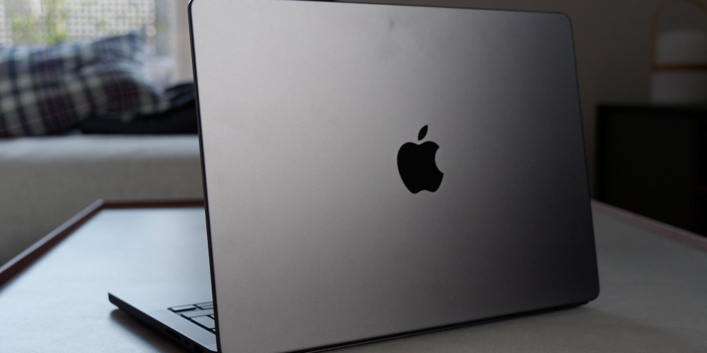 Imagen de un MacBook con el logo de Apple visible, en una mesa con un fondo desenfocado que incluye una cama y un sofá.