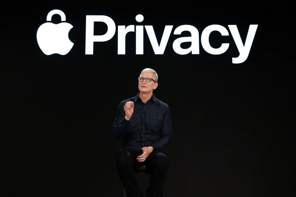 Un presentador de Apple sentado en una silla en un escenario oscuro, con un gran logotipo de Apple y la palabra 'Privacy' en el fondo.