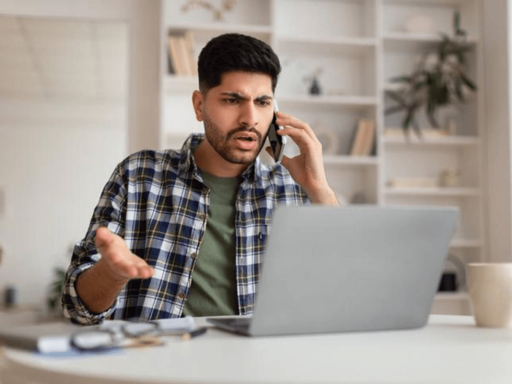 Un hombre con una camisa de cuadros está hablando por teléfono mientras mira con sorpresa su computadora portátil en un entorno de oficina.
