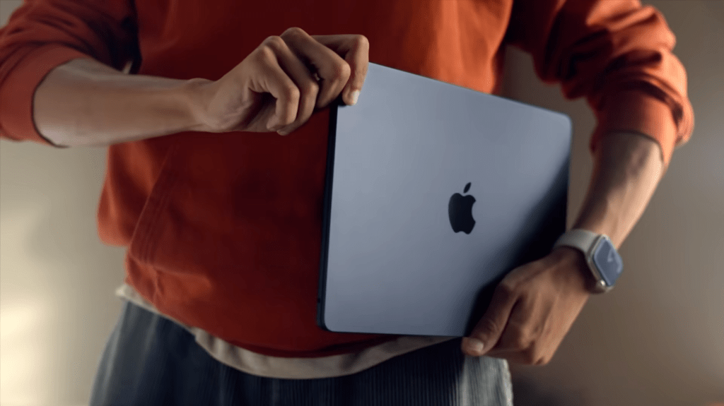 Persona sosteniendo un portátil Apple MacBook con una mano, mientras viste una sudadera naranja.