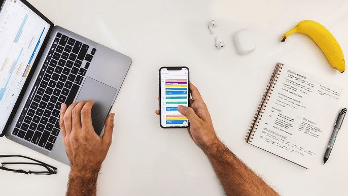 Una persona sostiene un teléfono móvil mostrando una aplicación de calendario, mientras trabaja en una computadora portátil. En la mesa hay un cuaderno con notas, un bolígrafo, un plátano y unos auriculares.