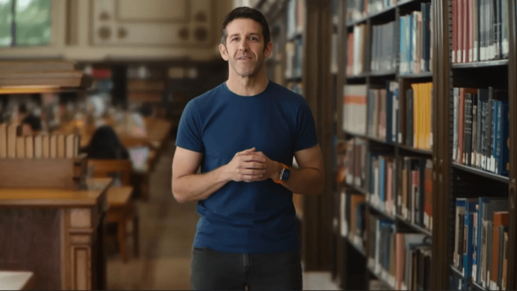 Un hombre de pie en una biblioteca, vestido con una camiseta azul y usando un reloj inteligente, mientras se comunica con la audiencia.