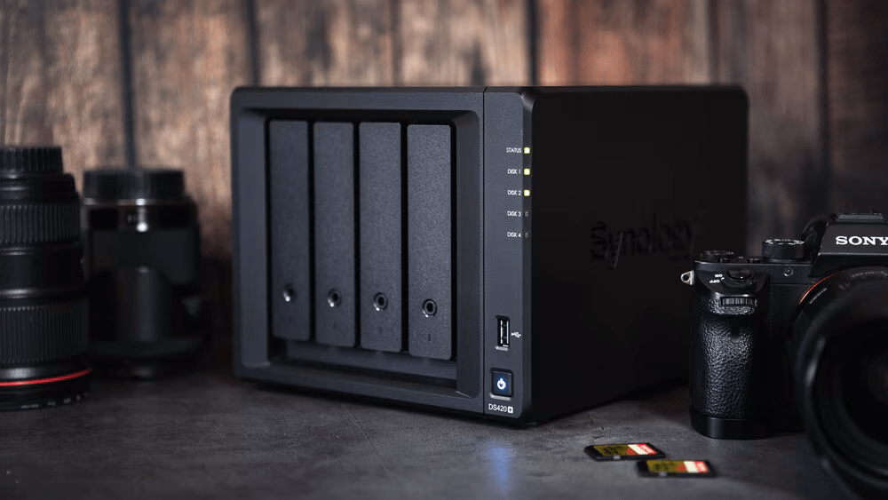 Un dispositivo NAS de Synology con cinco bandejas para discos duros, rodeado de cámaras y tarjetas de memoria en una mesa.