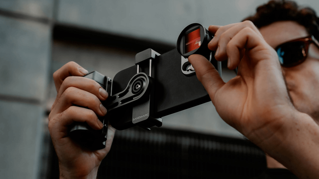 Una persona sostiene un soporte para teléfono inteligente, mientras ajusta un filtro en la lente del dispositivo, con un fondo que sugiere un entorno urbano.