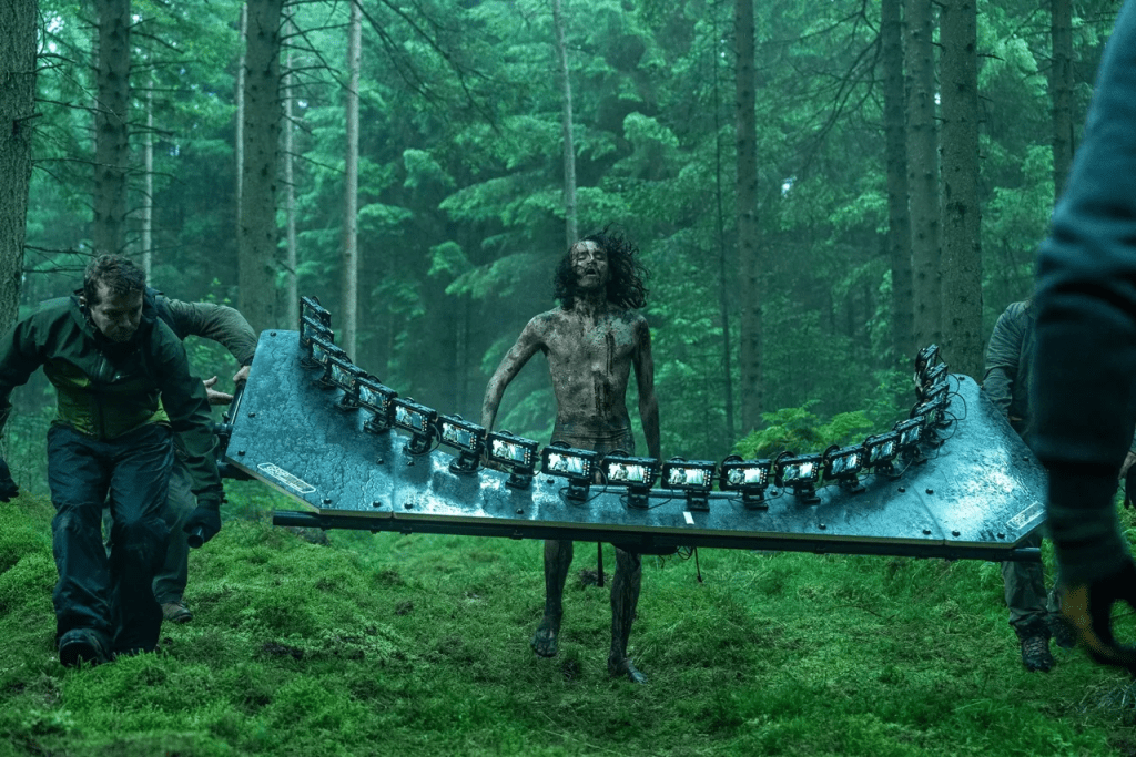 Un grupo de personas transporta un equipo de filmación por un bosque denso, mientras una persona lleva un rig con luces o cámaras montadas.