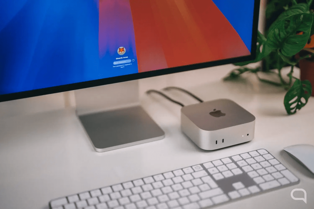 Un ordenador Apple Mac mini sobre un escritorio claro, junto a un teclado y un monitor que muestra la pantalla de inicio de sesión.