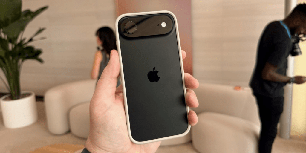 Imagen del iPhone Air 2 en la mano de una persona, mostrando su diseño elegante y ultrafino con una doble cámara trasera, fondo desenfocado con personas y muebles.