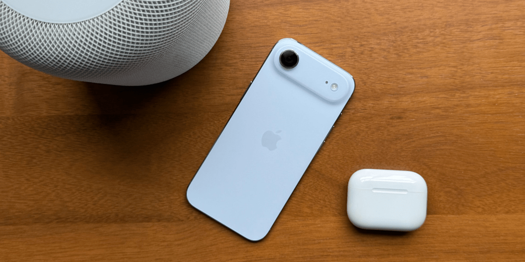 Imagen de un iPhone en color azul claro colocado sobre una mesa de madera, junto a un altavoz de Apple y unos AirPods en su estuche.