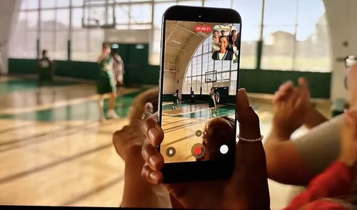 Una persona sostiene un teléfono móvil grabando un video en un gimnasio, mostrando a un jugador de baloncesto en acción.