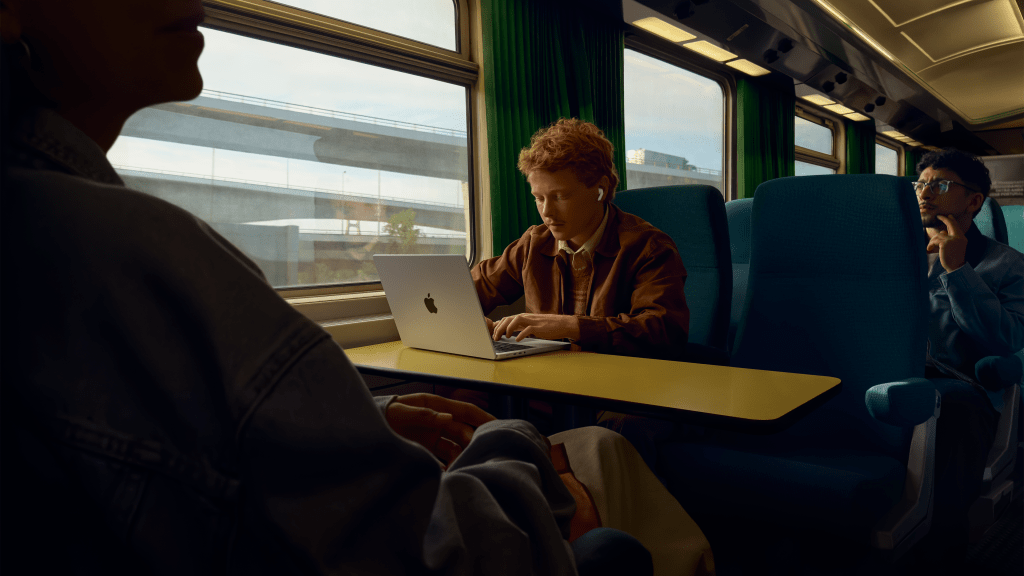 Una persona utilizando una laptop en un tren, con AirPods en sus oídos. A su lado, otra persona está sentada y mirando hacia la ventana, creando un ambiente de viaje moderno.