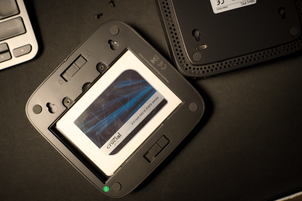 Vista superior de un mini PC con una unidad de almacenamiento SSD Crucial de 2.5 pulgadas colocada en su interior, sobre una superficie oscura.