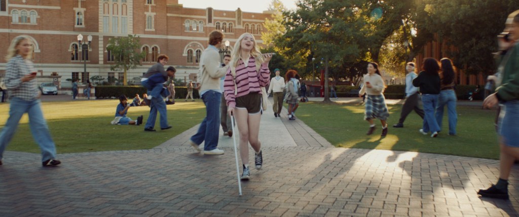 Estudiante caminando por el campus universitario con un bastón blanco, rodeada de otros estudiantes, con un ambiente soleado y árboles al fondo.