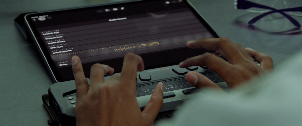 Manos utilizando un dispositivo de accesibilidad braille, mientras se muestra una pantalla de iPad con opciones de accesibilidad.
