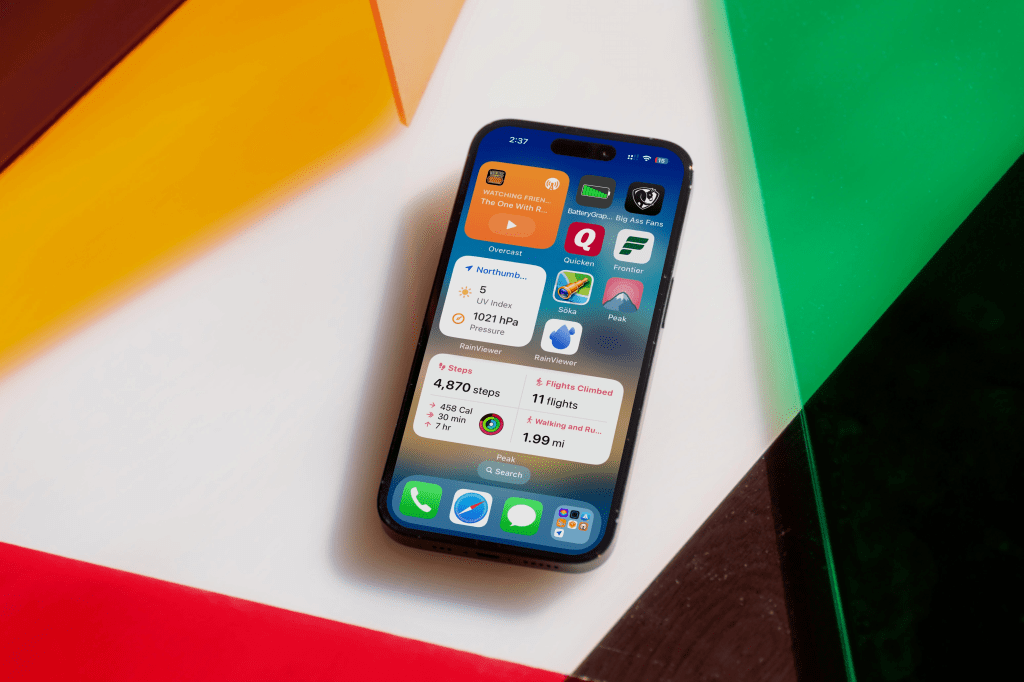 Un iPhone sobre una superficie con colores brillantes, mostrando la pantalla de inicio con varios widgets y aplicaciones organizadas.