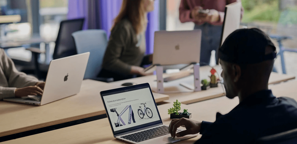 Grupo de personas trabajando en un espacio de oficina moderna, utilizando laptops Mac. Un individuo se enfoca en su computadora, que muestra una presentación de bicicletas, mientras otros interactúan en el fondo.