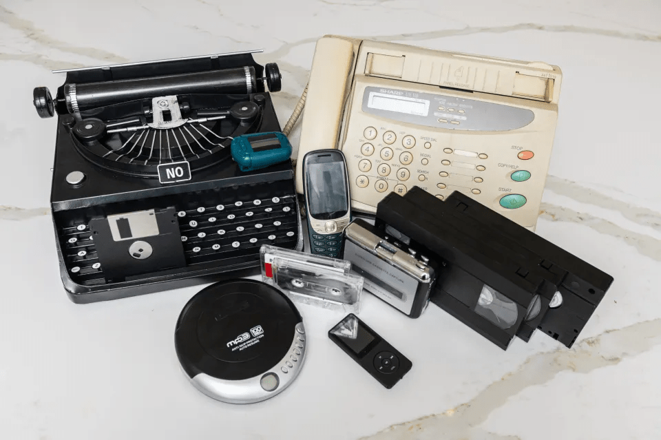 Una colección de dispositivos antiguos y obsoletos, incluyendo una máquina de escribir, un fax, teléfonos móviles, una grabadora de cassette, un reproductor de MP3 y un cassette de video, mostrando la evolución de la tecnología.