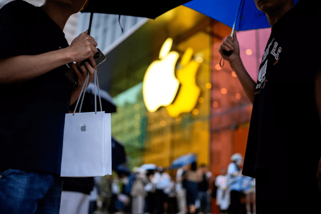 Two people holding umbrellas in front of an illuminated Apple store, one carrying a white Apple shopping bag.