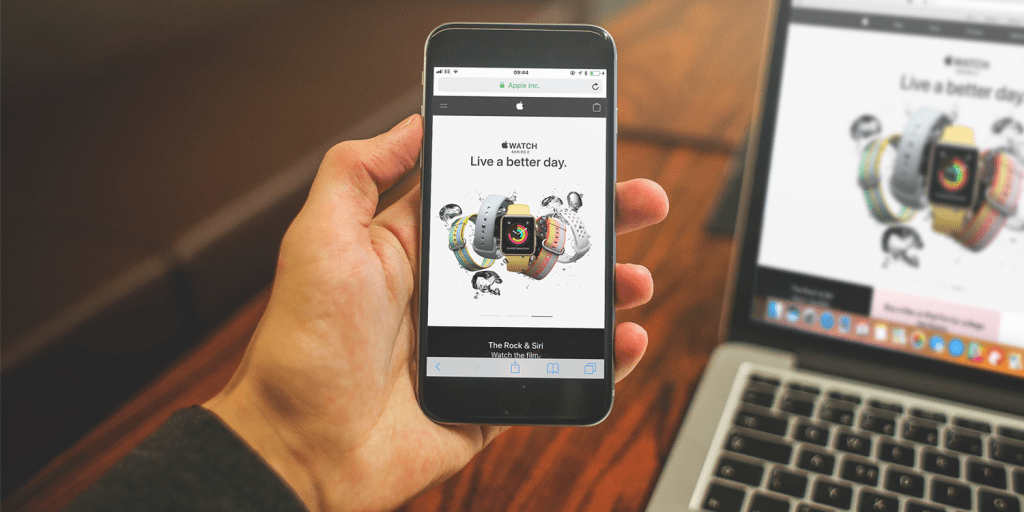 Una persona sosteniendo un iPhone que muestra la página web de un Apple Watch, mientras que un MacBook está al fondo mostrando la misma página.
