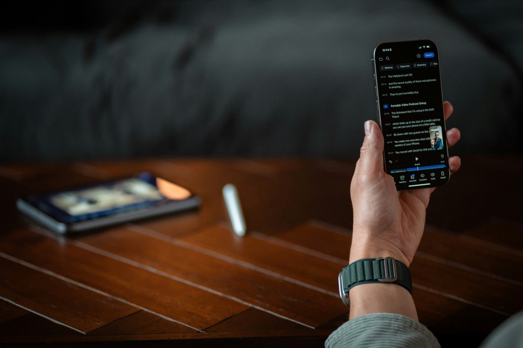 Una persona sosteniendo un iPhone en una mano, mostrando la pantalla con contenido. En el fondo, se ve otro dispositivo en una mesa de madera.