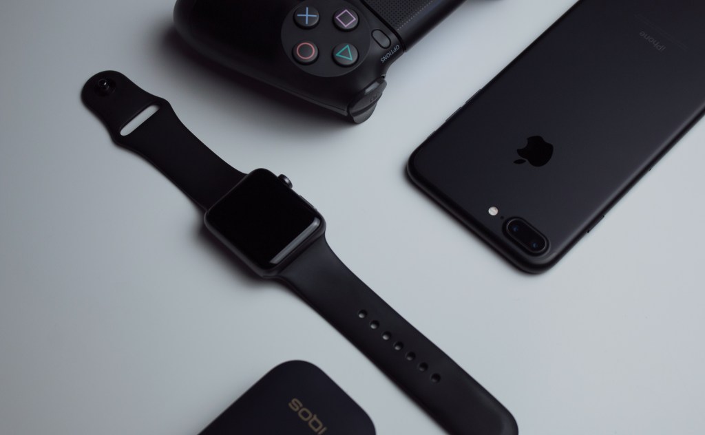 Imagen que muestra un reloj inteligente de Apple, un teléfono iPhone negro, un control de consola de videojuegos y un cargador en una superficie clara.