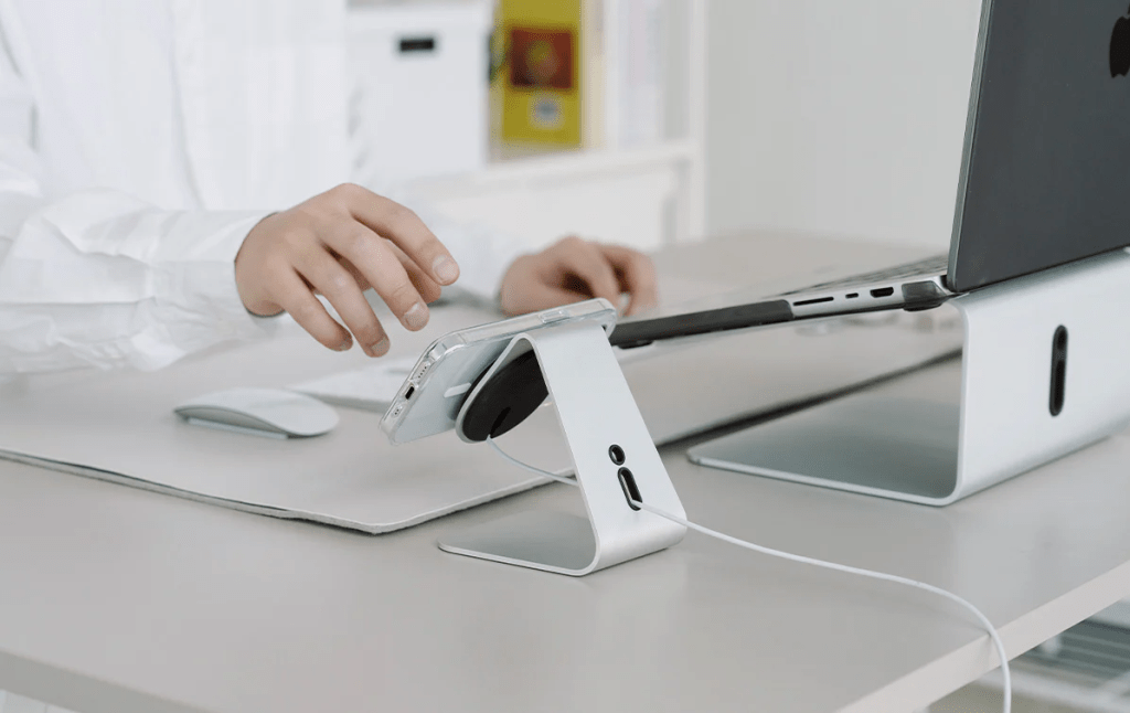 Una persona utilizando un soporte para teléfono móvil en un escritorio, con una computadora portátil y un mouse en el fondo.