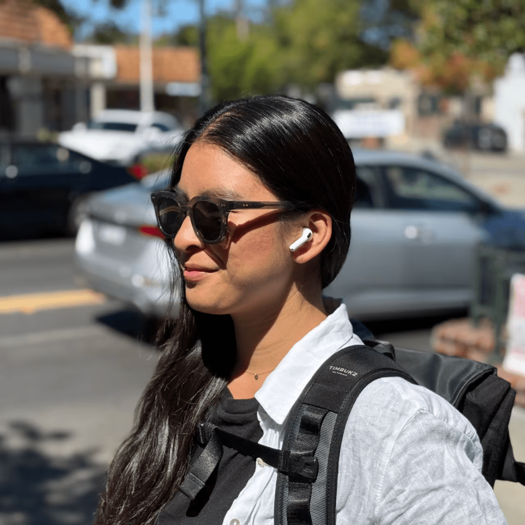 Mujer joven con gafas de sol y auriculares inalámbricos, sonriendo mientras camina por la calle, con una mochila en la espalda.