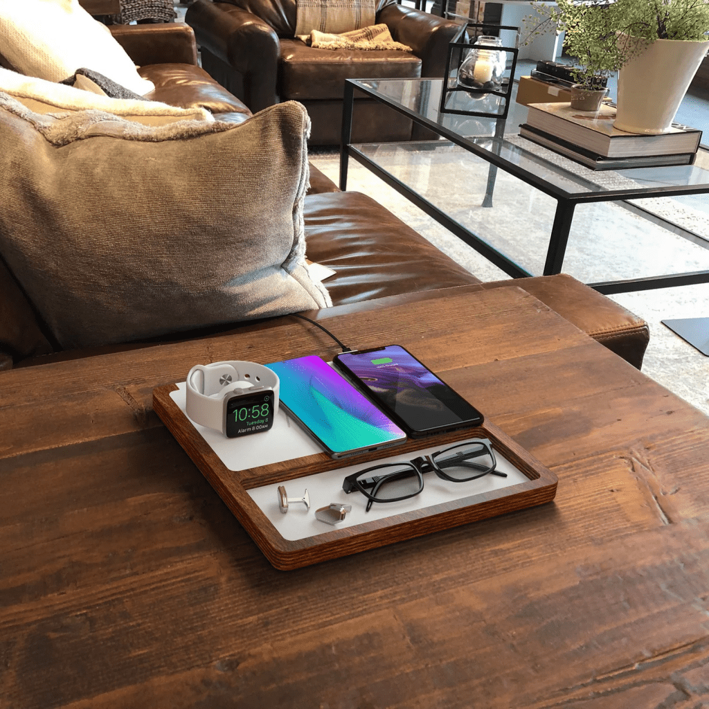 Una bandeja de madera con un reloj inteligente, un teléfono móvil, gafas, un par de audífonos y un sujetapapeles, todo sobre una mesa de madera junto a un sofá.
