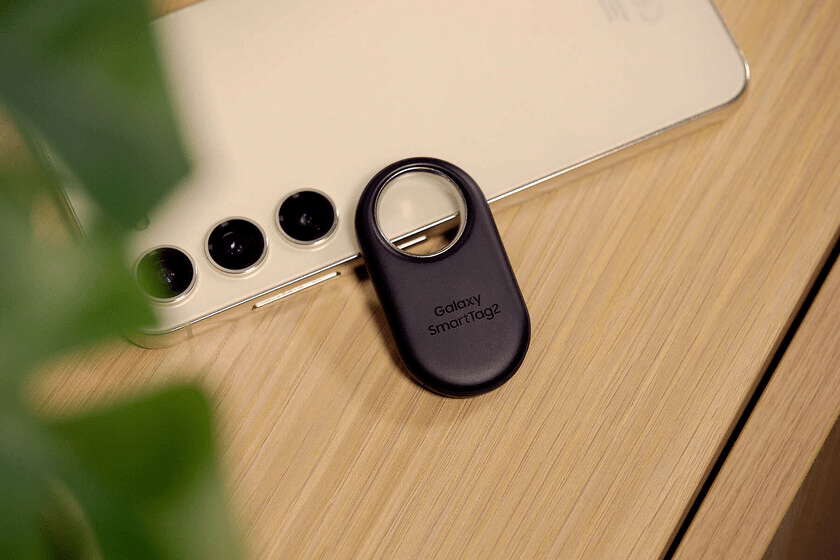 Un Galaxy SmartTag2 negro ubicado sobre un teléfono móvil de color dorado en una superficie de madera.