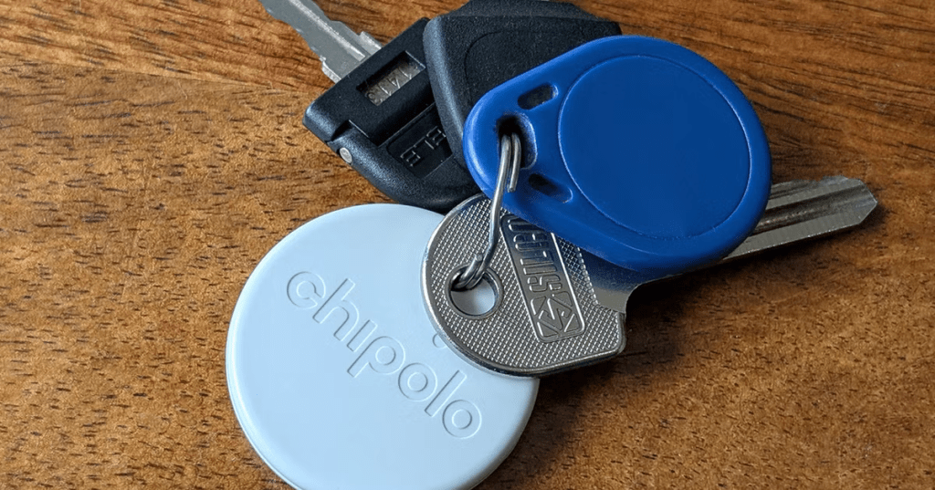 Un conjunto de llaves sobre una superficie de madera, incluyendo un llavero Chipolo de color blanco, una llave de metal y un objeto azul en forma de disco.
