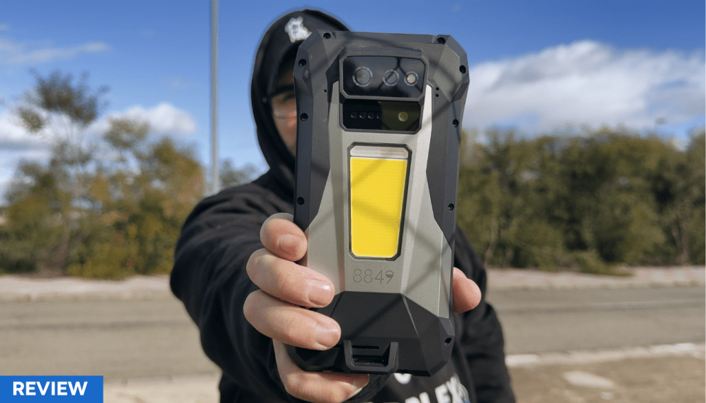 Una persona sostiene un teléfono móvil robusto y moderno con una carcasa protectora, frente a un paisaje natural y un cielo despejado.