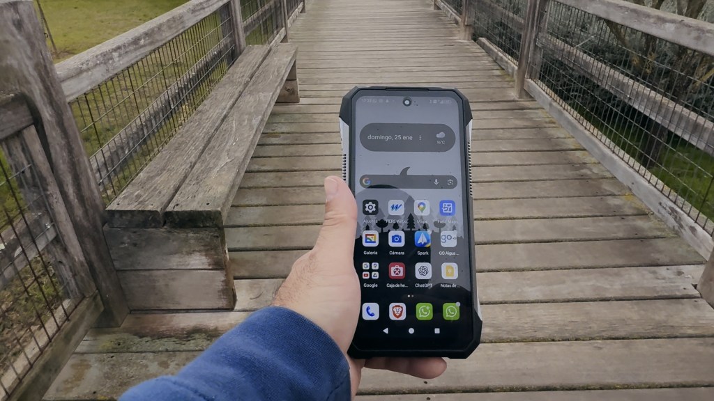 Un primer plano de un teléfono móvil en una mano, mostrando su pantalla con aplicaciones, mientras está sostenido sobre un sendero de madera con bancos a los lados.