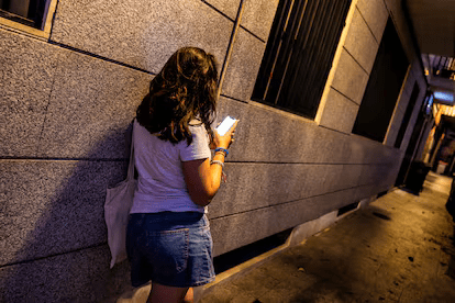 Una persona de espaldas, con cabello rizado, está de pie junto a una pared gris, usando su teléfono móvil. La escena tiene iluminación tenue y un ambiente urbano.