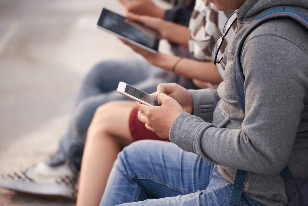 Grupo de jóvenes sentados, cada uno usando un dispositivo móvil, como teléfonos y tabletas, en un entorno al aire libre.