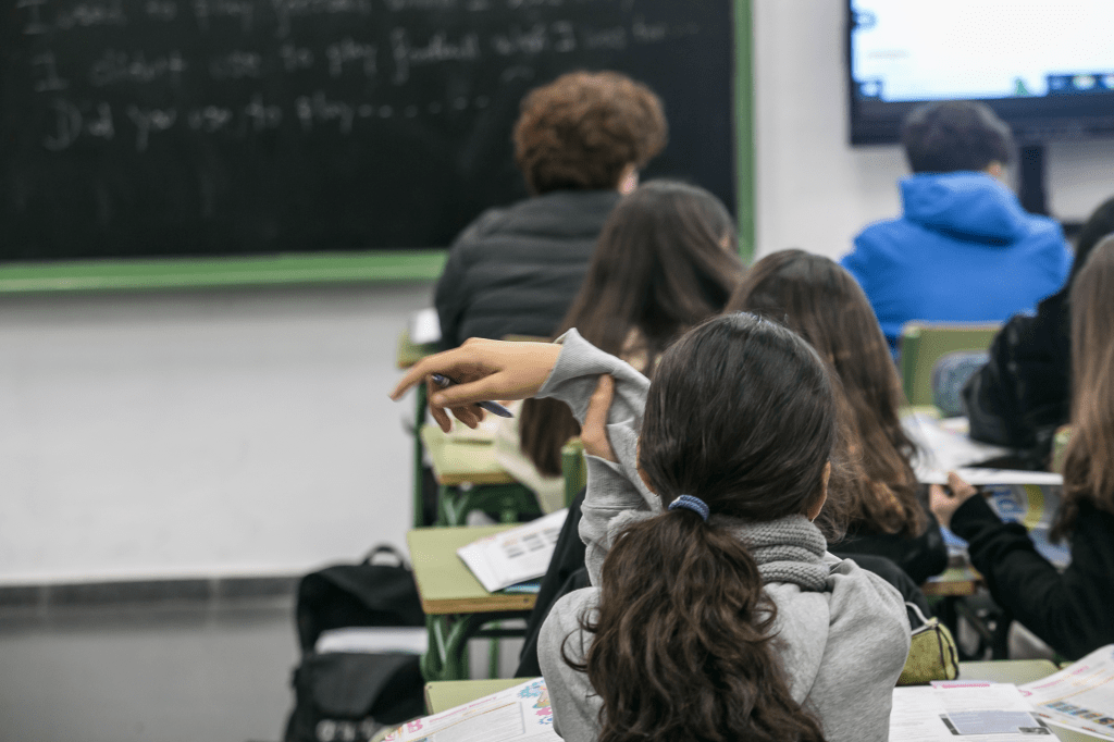 Estudiantes en un aula con la atención en el profesor, una niña levanta la mano para participar.