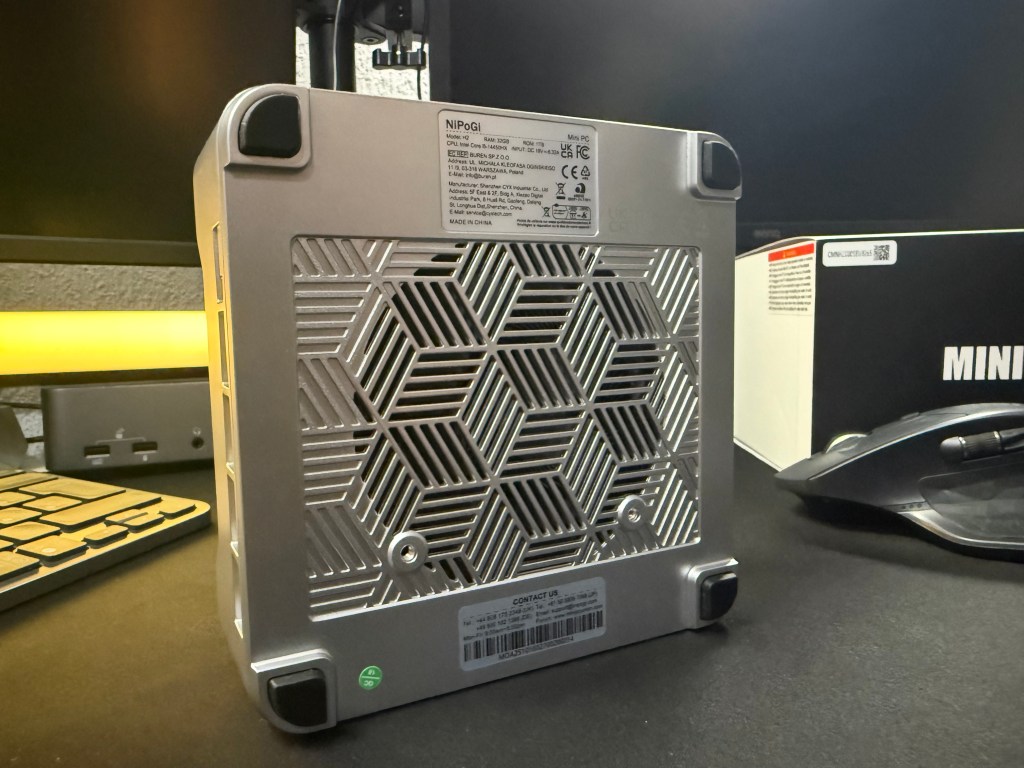 Vista lateral de un mini PC NipoGi con un diseño de ventilación en forma de panal, mostrando la parte trasera con etiquetas y conexiones, sobre un escritorio oscuro con un teclado y mouse.