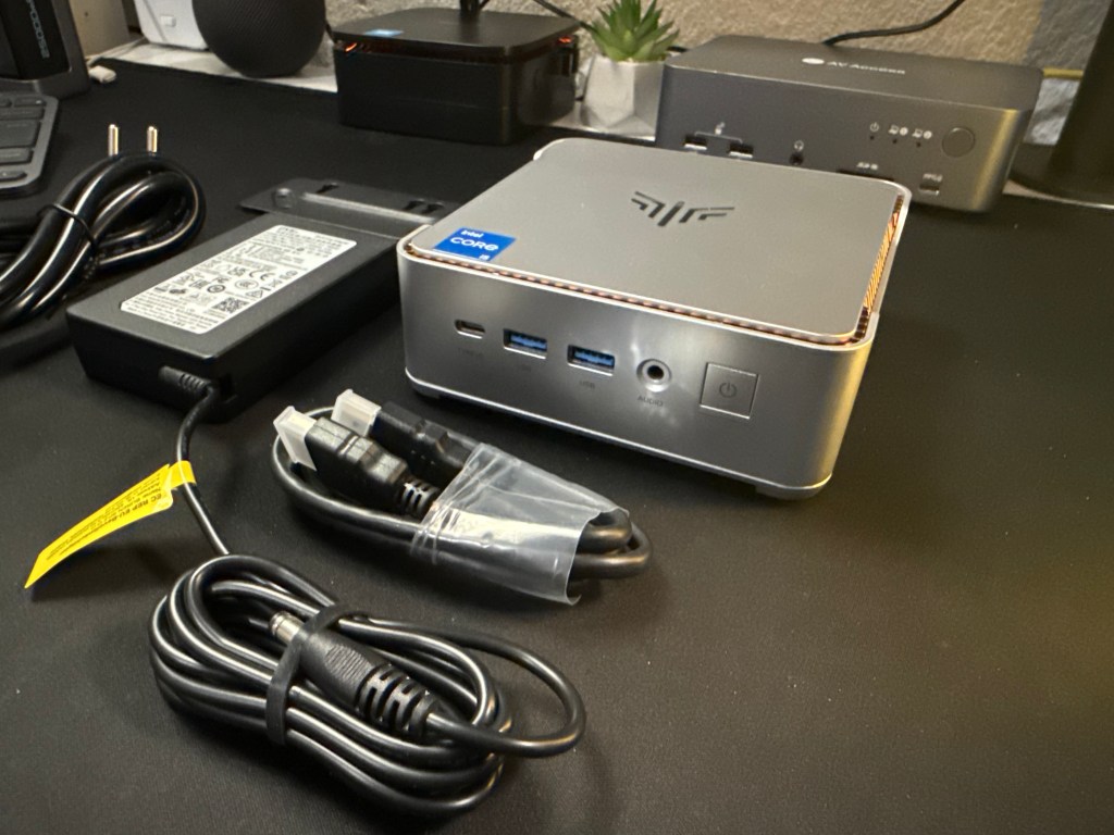 Mini computadora con puertos USB y de audio, acompañada de cables y adaptador de corriente.